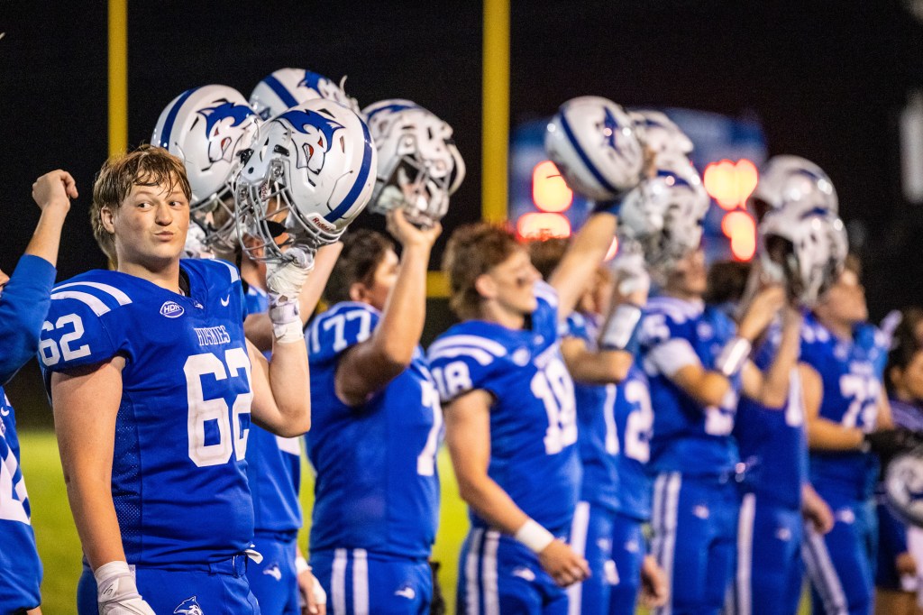 A group of football players in blue uniforms hold their helmets high, celebrating on the sidelines during a game.