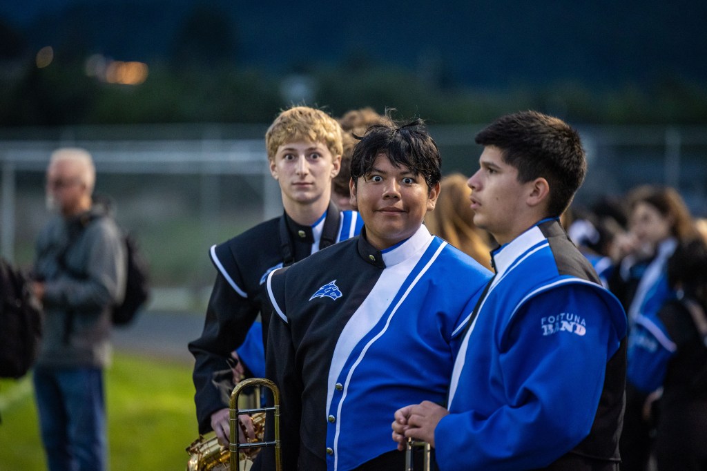A group of band students in uniforms stand outside, with one student looking surprised and holding a musical instrument. The background shows a blurred crowd and evening lighting.