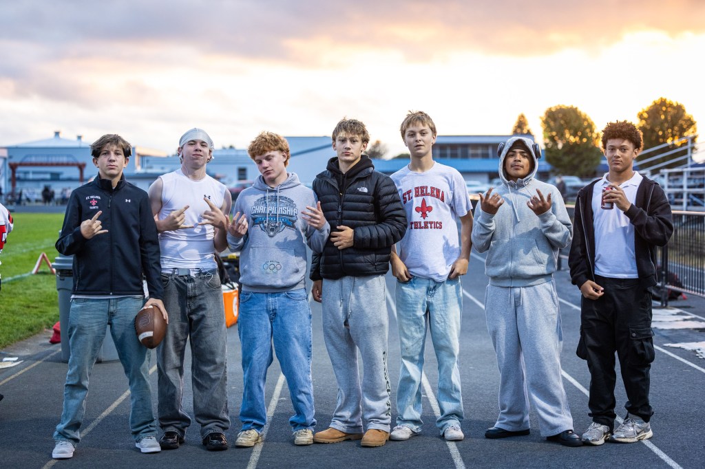 A group of seven young men standing together on a track field, posing casually. Some are wearing athletic gear and relaxed clothing. The sky in the background shows a sunset with soft clouds.