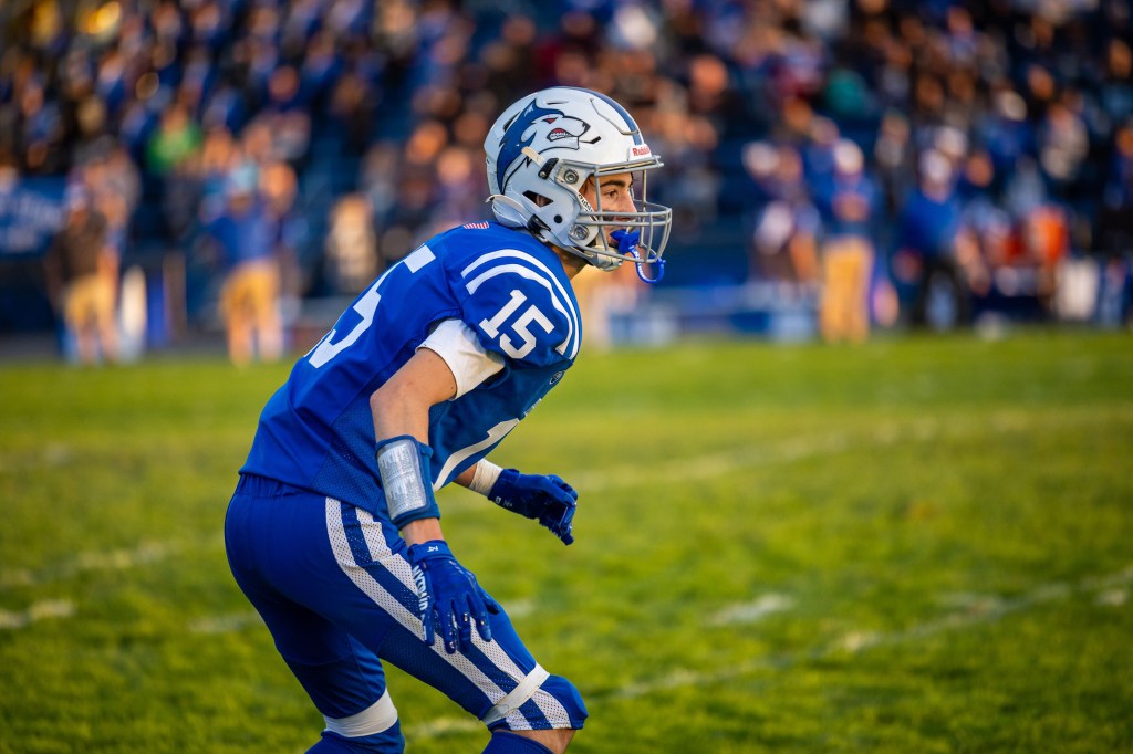 Football player in blue uniform number 15, preparing for a play on the field with spectators in the background.