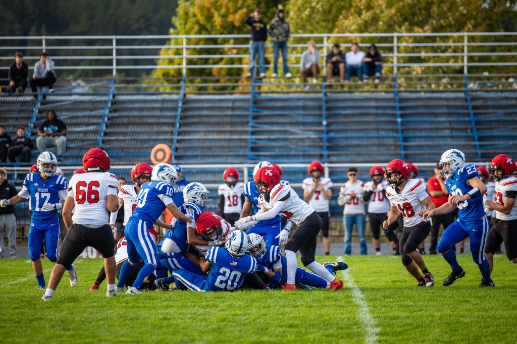 A football game in action with players from two teams in the midst of a tackle on the grassy field, spectators in the background on bleachers.