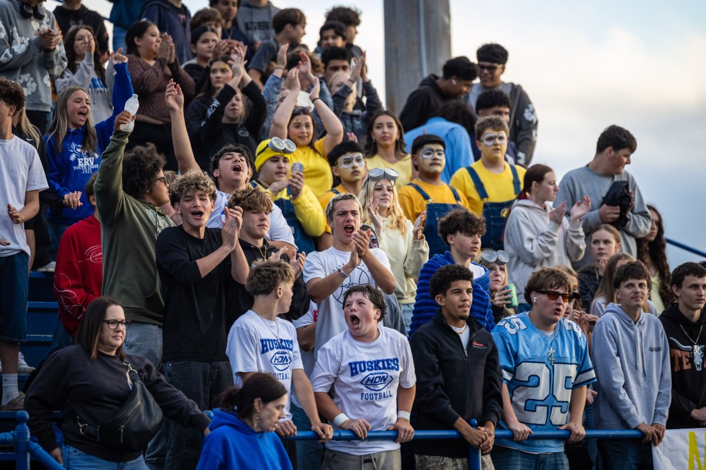 A large group of enthusiastic high school students cheering and celebrating at a football game, some wearing school apparel and face paint.