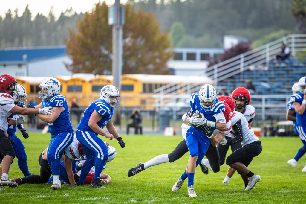 A high school football game in progress, with players in blue and red jerseys competing on a grassy field. A player in blue is running with the ball while being tackled by multiple defenders.
