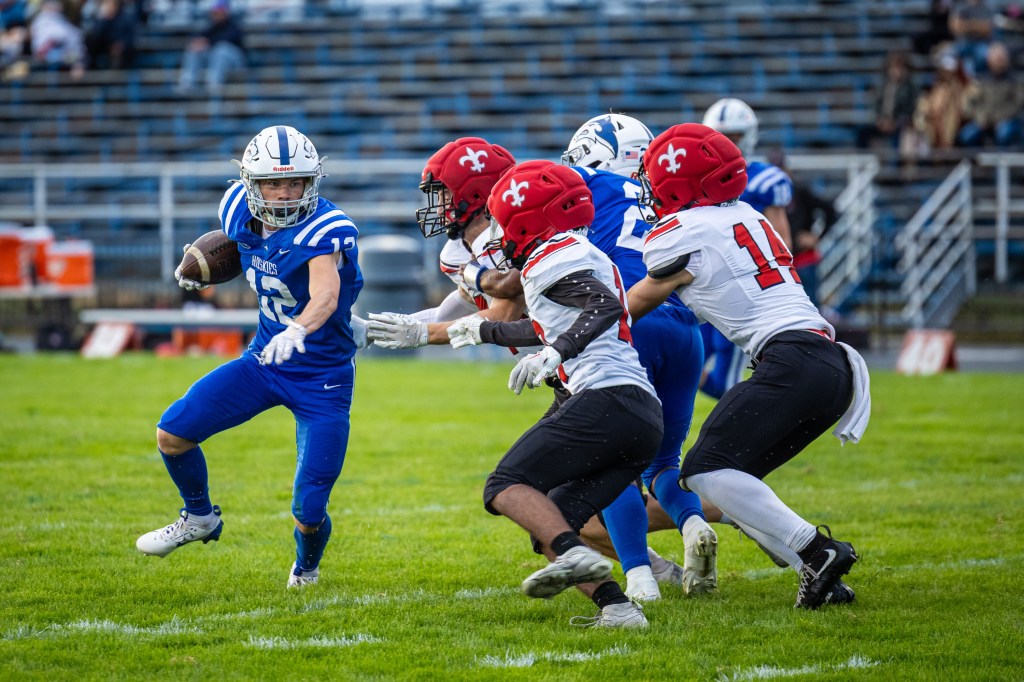 A high school football player in blue uniform evades defenders in red and white uniforms during a game on a grassy field.