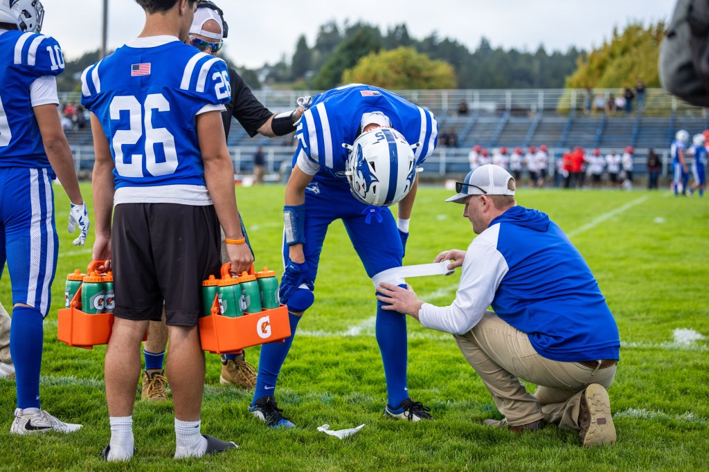 A football player in blue uniform is being attended to by a coach on the field, while teammates and water bottles are visible in the background.