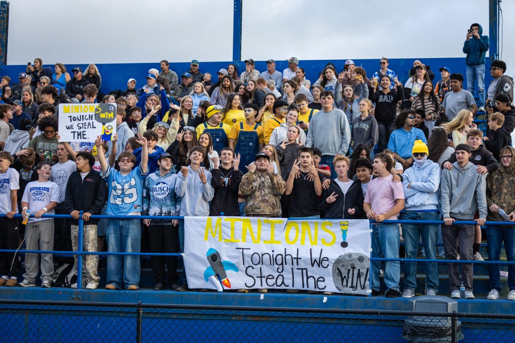 A large crowd of enthusiastic students at a school event, holding signs that say 'Minions' and 'Tonight we steal the win'. The students are wearing various team colors and some are dressed in themed outfits.