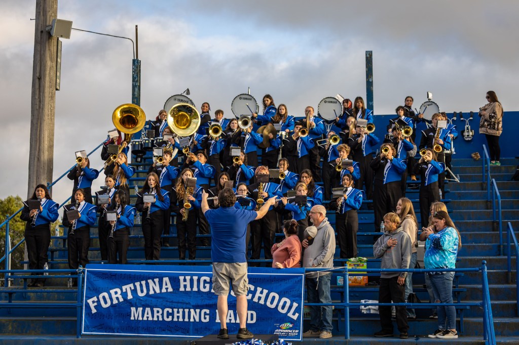 A marching band in blue uniforms performs on bleachers, with various instruments visible, including brass and woodwinds. A conductor is leading the band while an audience watches from below.
