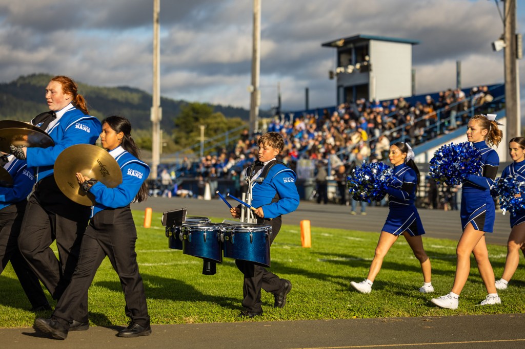 A marching band and cheerleaders performing at a football game, with musicians carrying instruments and cheerleaders holding pom-poms, set against a backdrop of an enthusiastic crowd in the stands.