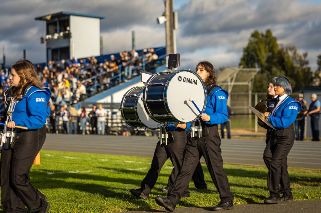 A marching band performs on a sports field, with musicians dressed in blue uniforms. One member is carrying a large bass drum labeled 'Yamaha'. The audience can be seen in the background.