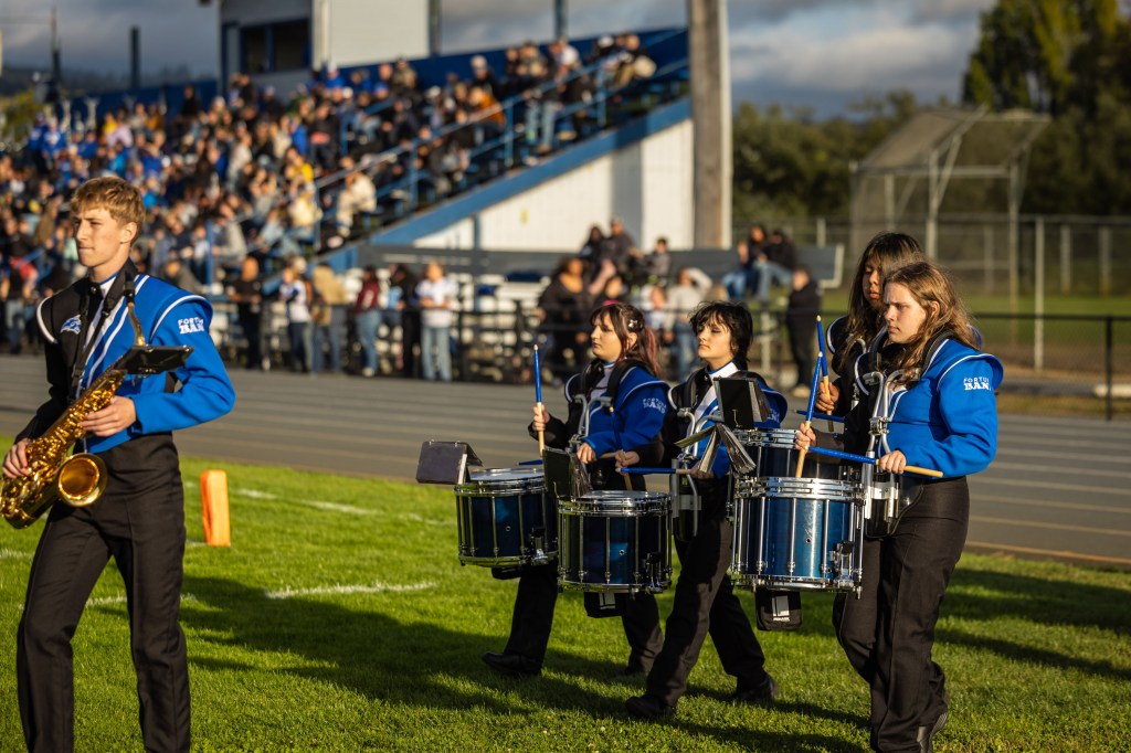 A marching band performing on a grassy field, featuring a saxophonist in a black and blue uniform and several drummers in matching attire.