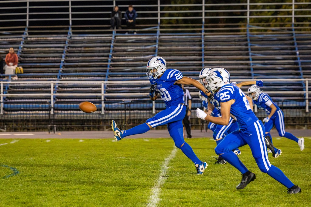 A football player in a blue uniform kicks a football on a grassy field, with teammates in blue uniforms running nearby and empty bleachers in the background.