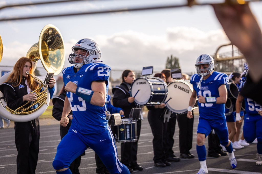 A group of football players in blue uniforms runs onto the field, accompanied by a marching band playing instruments. The focus is on player number 23, showing determination as they enter the field.