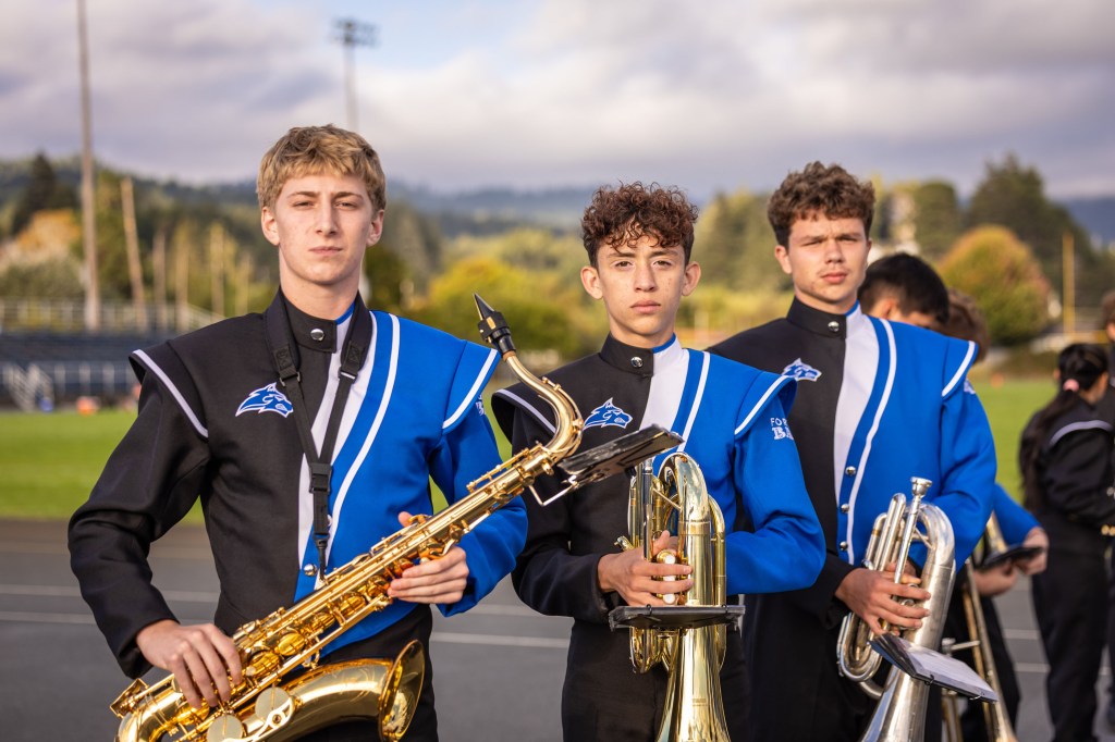 Three young musicians in marching band uniforms holding musical instruments, including a saxophone and brass instruments, standing on a field with trees in the background.