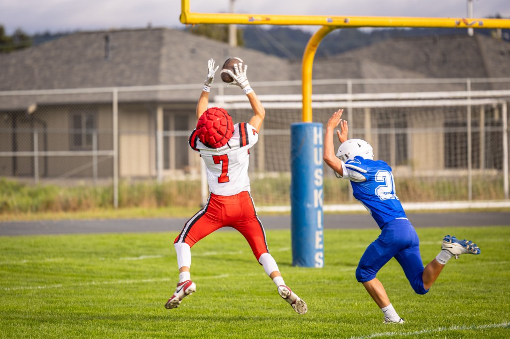 A football player in a red uniform with the number 7 leaps to catch a football above his head, while a player in a blue uniform with the number 22 attempts to defend against him.