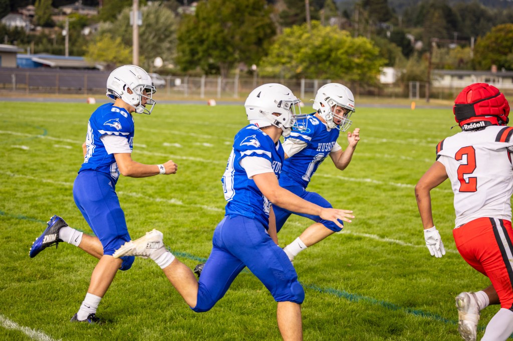 Three football players in blue jerseys chase an opposing player in a red jersey on a grassy field during a game.