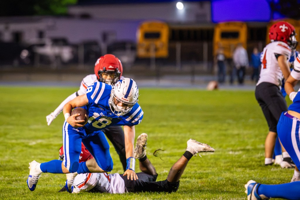 A high school football player in blue and white uniform dodges a tackle while running on a grassy field during a night game.