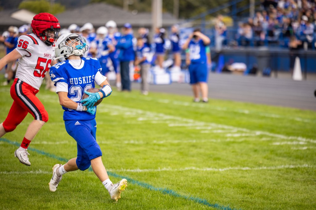 A youth football player in a blue Huskies jersey runs on the field while a player in a red jersey approaches from behind during a game.