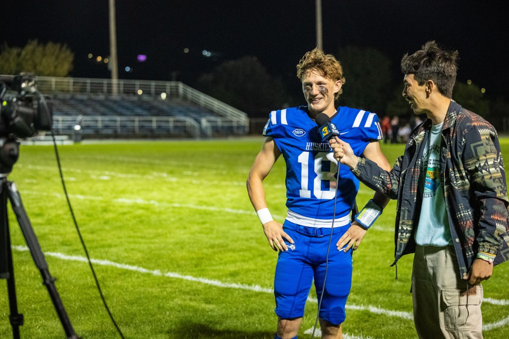 A young football player wearing a blue jersey with the number 18 is being interviewed on a field at night. He is smiling while a reporter, dressed in casual attire, holds a microphone and asks questions. In the background, there are empty bleachers and stadium lights.