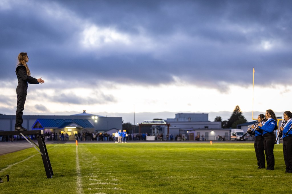 A conductor stands on a podium directing a marching band at a football field during twilight.