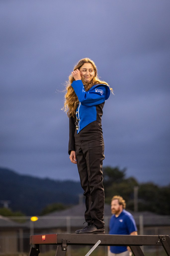 A young person with long hair stands confidently on a platform, wearing a blue and black outfit, looking cheerful against a cloudy sky.