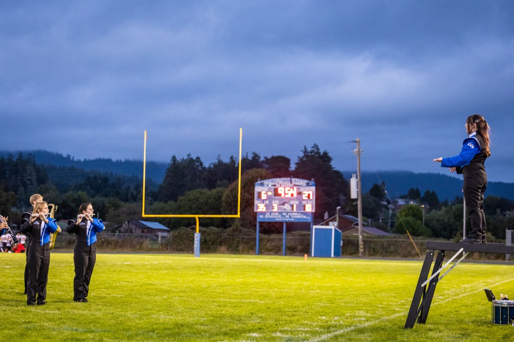 A band performing on a football field at dusk, with musicians playing woodwind instruments and a conductor standing on a platform, while a scoreboard shows the game time and score.