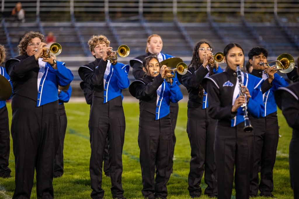 A group of young musicians in blue and black uniforms playing instruments at a football field during a performance.