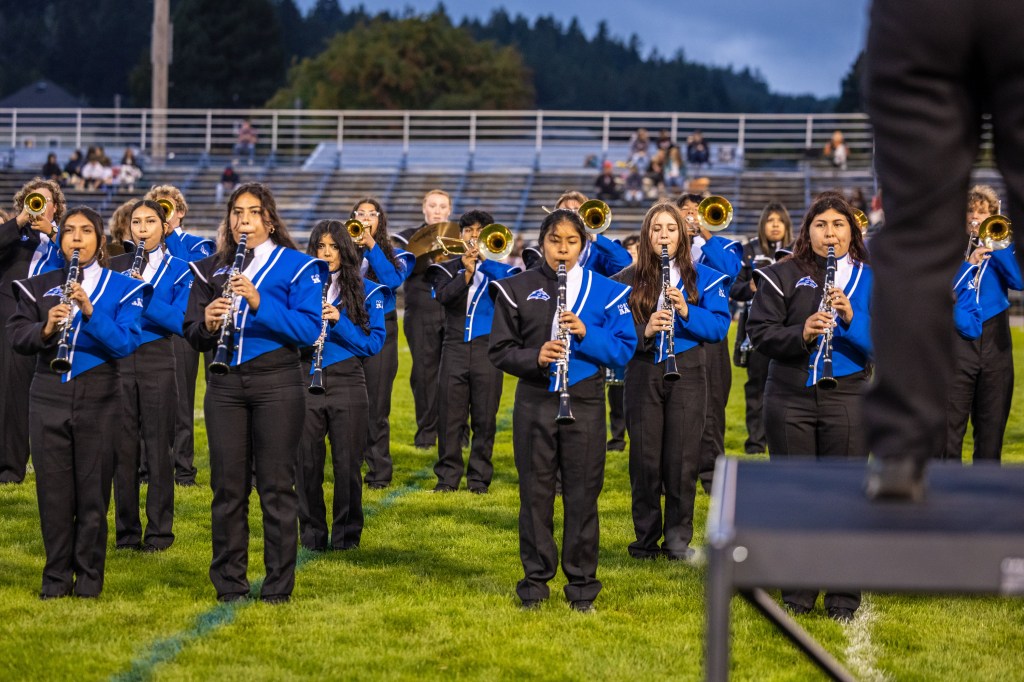 A marching band performing on a football field, featuring musicians playing clarinets and trumpets, wearing blue and black uniforms.