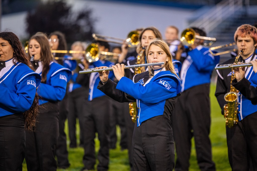 A marching band performing on a field, featuring a flutist in a blue and black uniform, surrounded by other band members playing brass instruments.