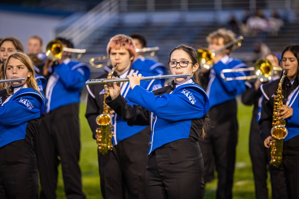 A group of marching band members performing, with a focus on a young female flutist wearing glasses and a blue uniform, surrounded by other musicians playing brass and woodwind instruments.