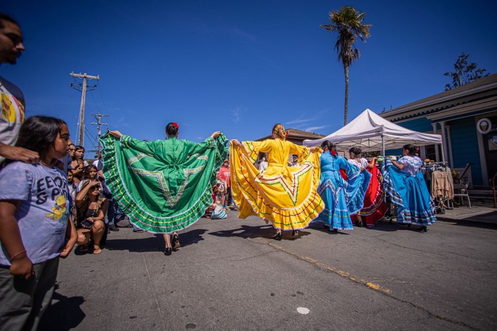 A group of dancers wearing colorful traditional dresses perform in a street festival, with an audience watching. The dancers' outfits include green, yellow, blue, and red dresses, while the backdrop features a clear blue sky and palm trees.