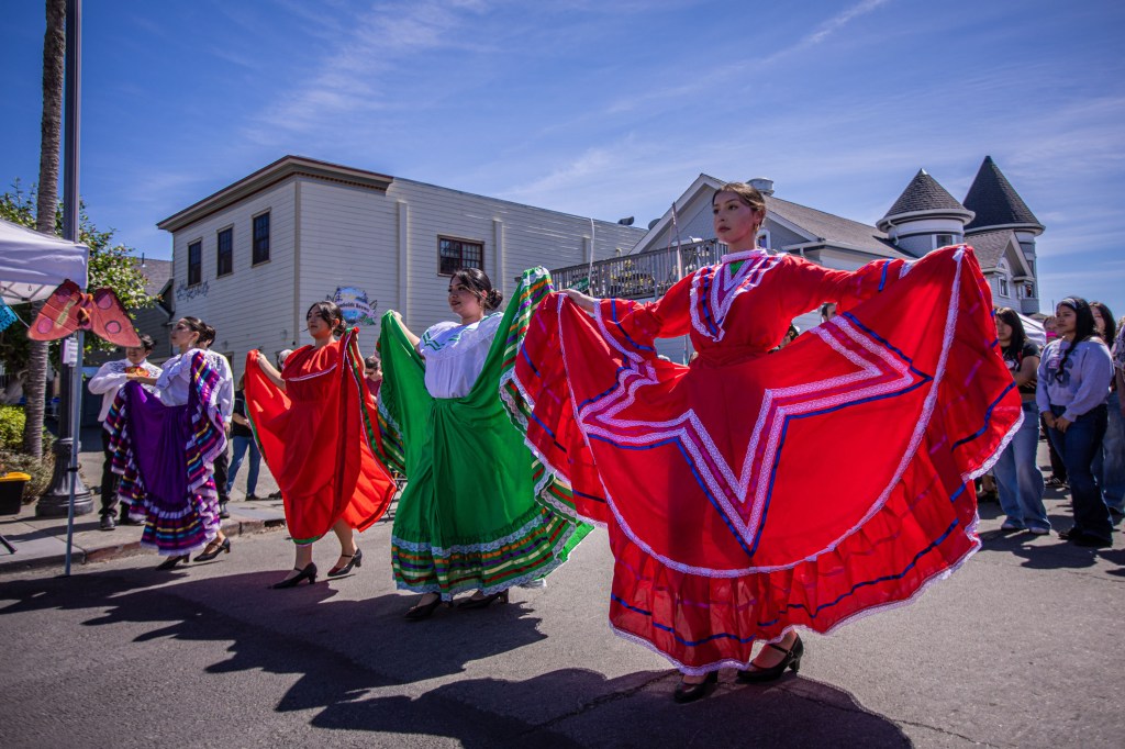 A group of dancers in traditional colorful dresses performing in a street festival, with a sunny sky and buildings in the background.