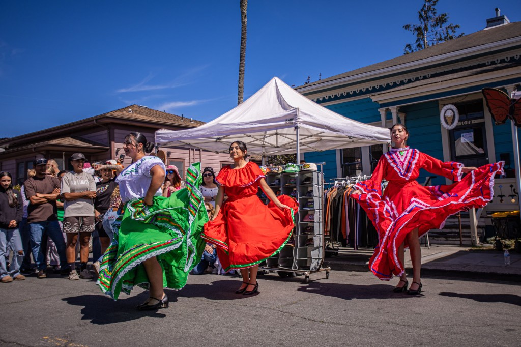 Three dancers performing a traditional cultural dance in colorful dresses at an outdoor event, with an audience watching in the background.