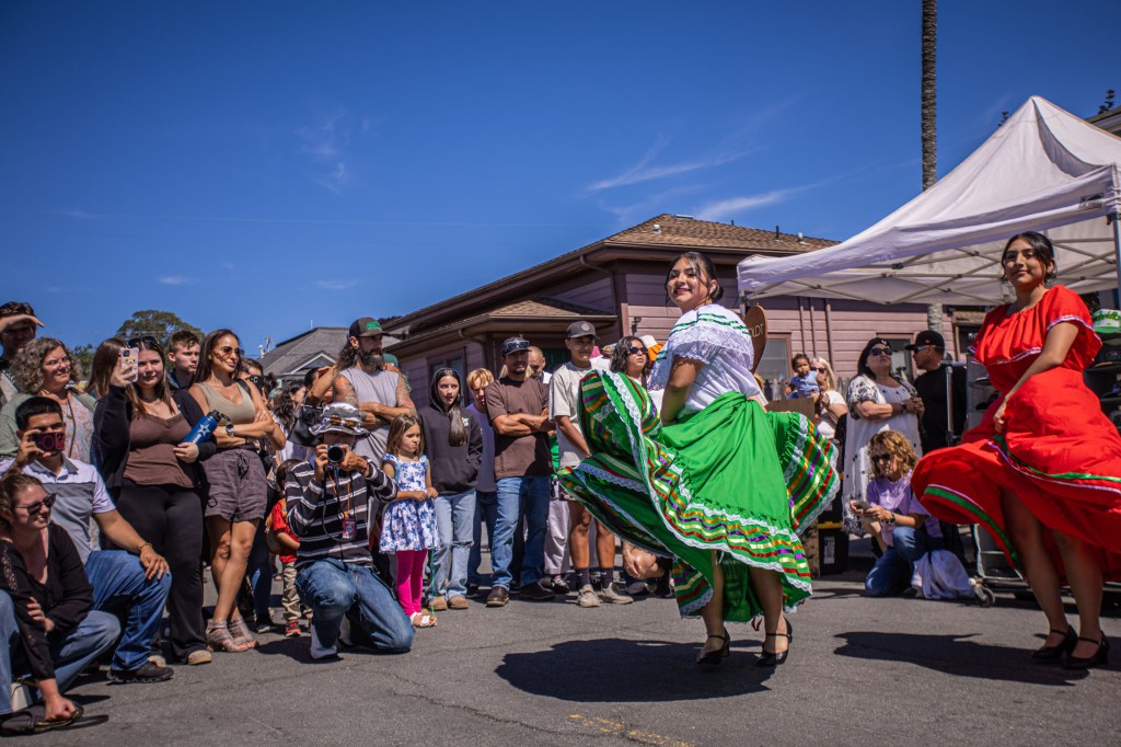 Two dancers in colorful traditional dresses perform on a street in front of an audience during a cultural festival, with a clear blue sky above.
