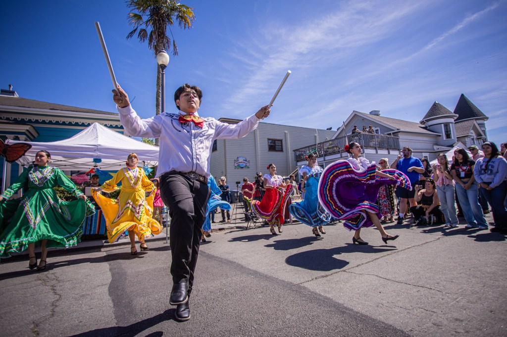 A group of dancers in colorful traditional outfits performing on a street during a cultural festival. A male dancer is leading the performance with a baton, while others spin in vibrant dresses.