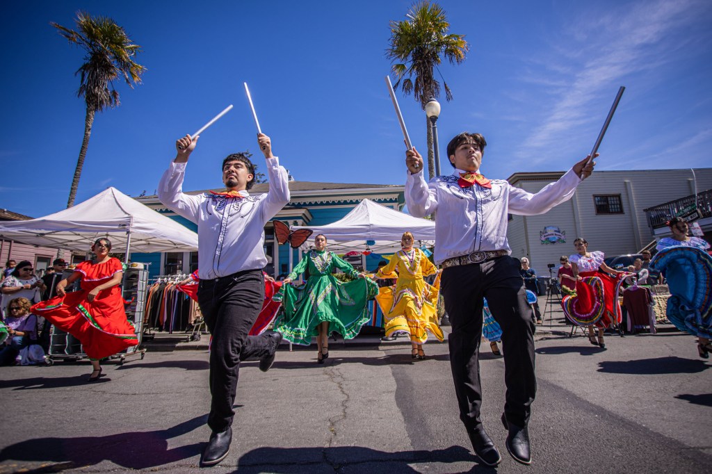 A vibrant cultural celebration featuring dancers in colorful traditional costumes performing in an outdoor setting, with palm trees and tents in the background.