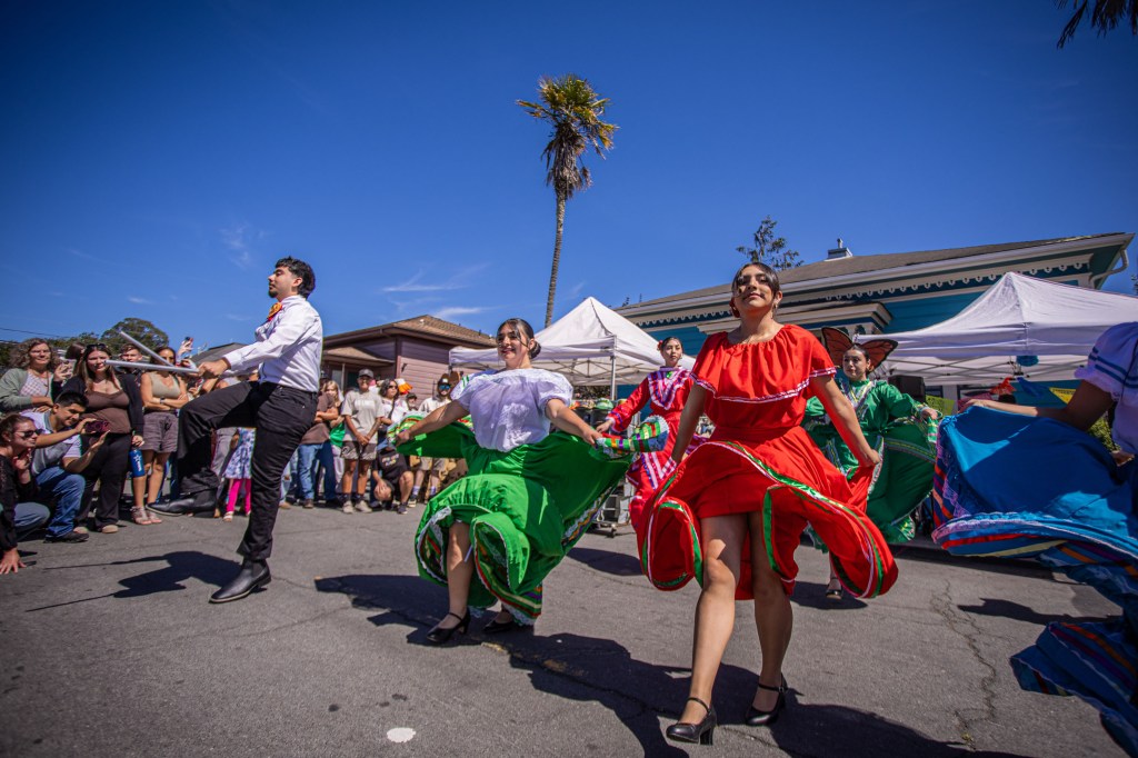 A group of dancers performing a traditional dance in colorful costumes during an outdoor event, surrounded by an engaged audience.