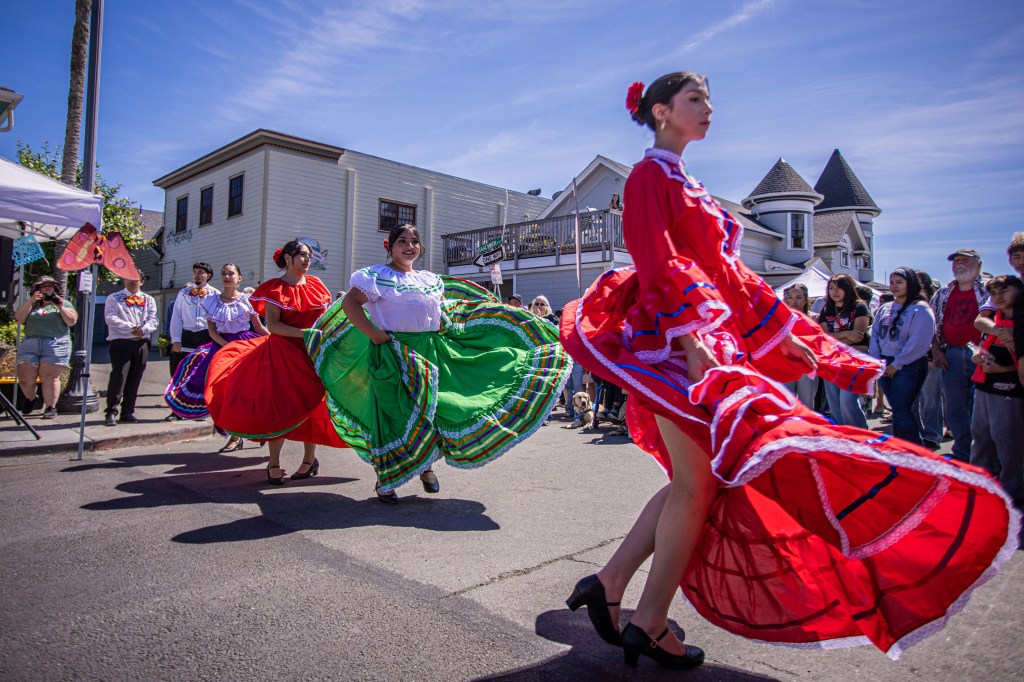A vibrant cultural dance performance featuring women in colorful traditional dresses, dancing in an outdoor setting with spectators watching.