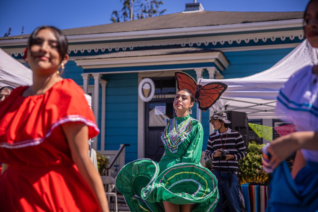 A dancer in a vibrant green traditional outfit with a butterfly headpiece performs at a cultural festival, surrounded by other performers in colorful attire, with a blue building and tents in the background.