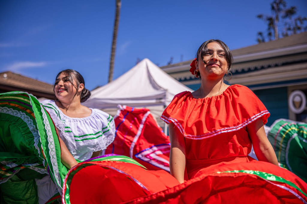 Two women dancing in traditional Mexican dresses, one in a red dress and the other in a white dress with green and red accents. They are performing at an outdoor event with tents in the background and a clear blue sky.