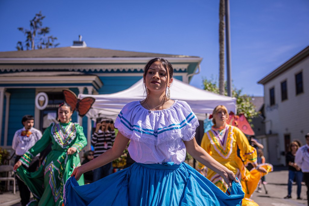 A group of dancers in colorful traditional attire performing in a lively outdoor setting, with a blue sky and a festive atmosphere.