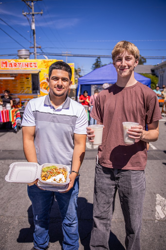 Two young men standing in a street fair, one holding a takeaway food container filled with nachos and the other holding two drinks. Colorful food trucks and festival tents in the background under a clear blue sky.