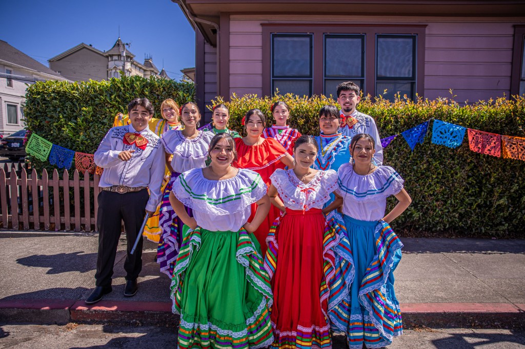 Group of individuals posing in traditional colorful outfits with a festive backdrop of papel picado decorations and a house.