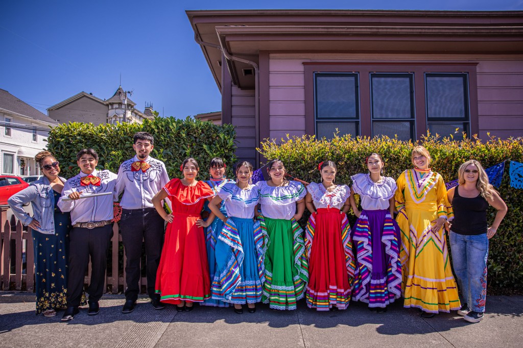 A group of people dressed in colorful traditional attire posing in front of a house with greenery in the background on a sunny day.