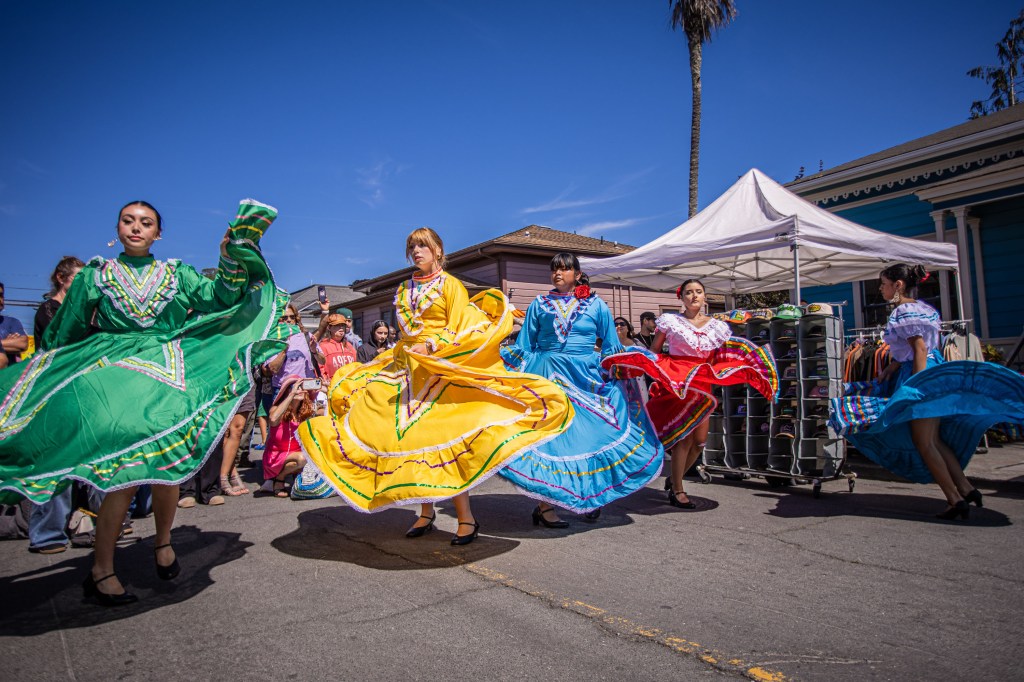 A group of women in colorful traditional dresses performing a cultural dance in a street festival, with spectators watching in the background.