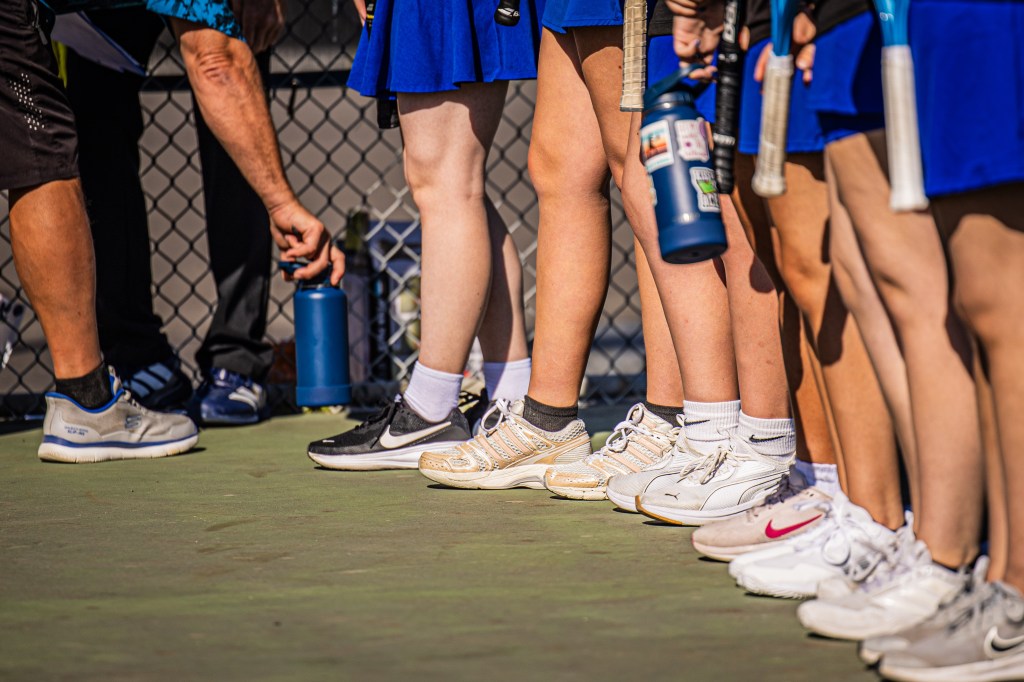 Close-up of multiple pairs of feet in tennis shoes on a court, with players in blue skirts and a coach in the background holding a water bottle.