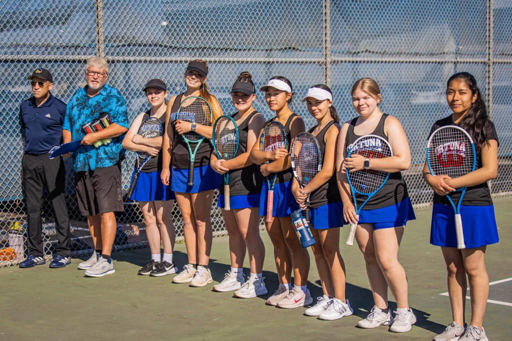 A group of eight young female tennis players on a court, standing in a line with their rackets. Two coaches are on the left, with one holding awards.