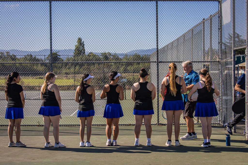 A group of female tennis players in black tops and blue skirts stand in a line, listening to a coach on a tennis court with a fence and mountains in the background.