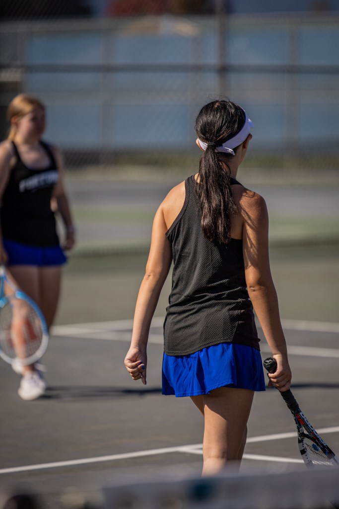 Two female tennis players on a court, one in a black top and blue skirt, facing away from the camera, holding a tennis racket.