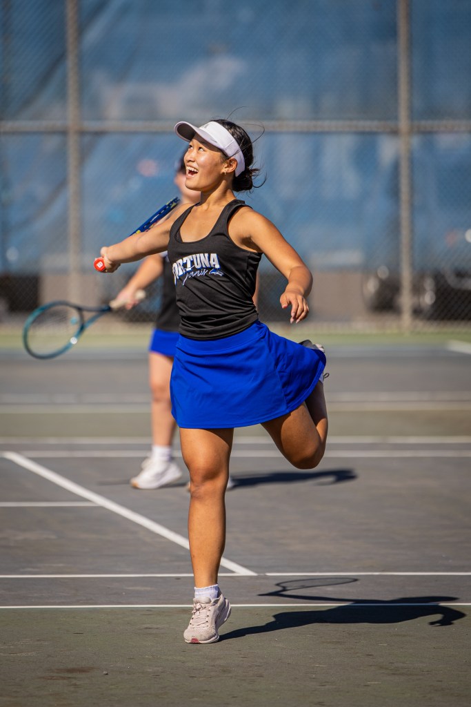 A young female tennis player in a black tank top and blue skirt joyfully running on a tennis court, holding a racket, with a smile on her face.