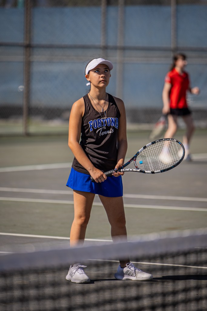 A young female tennis player in a black and blue outfit, holding a tennis racket, appears focused on the game while standing on the court. A blurred figure in a red shirt can be seen in the background.
